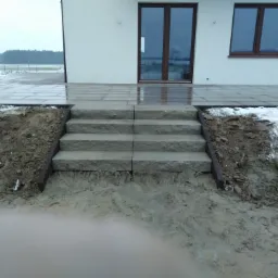 Kamienne schody prowadzące na taras z płytek brukowych przy białym domu w pochmurny dzień.