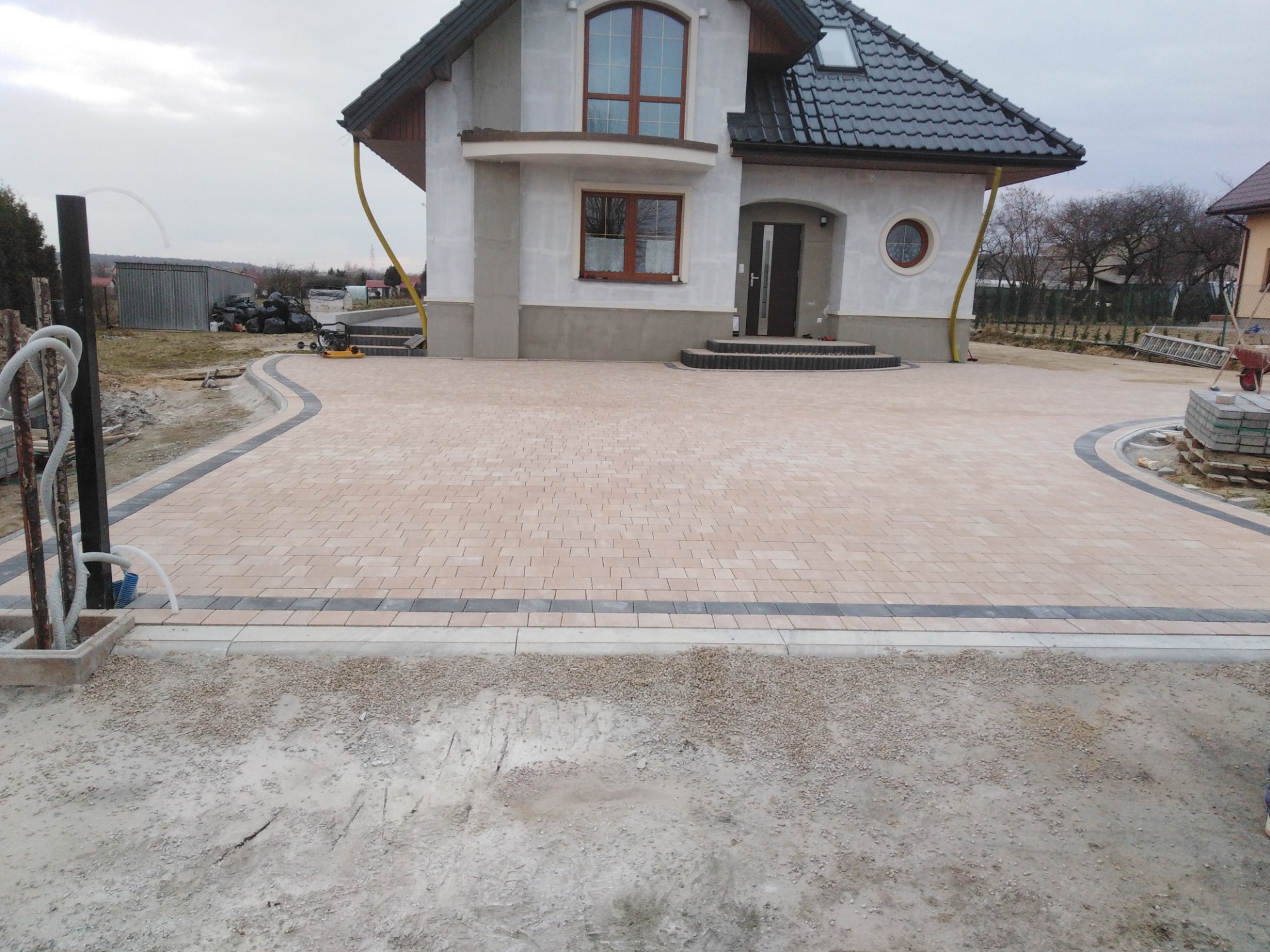 Nowo ułożona beżowa kostka brukowa z ciemnoszarą obwódką przed domem z szarą elewacją, widoczne przygotowania do dalszych prac wykończeniowych i elementy instalacji.