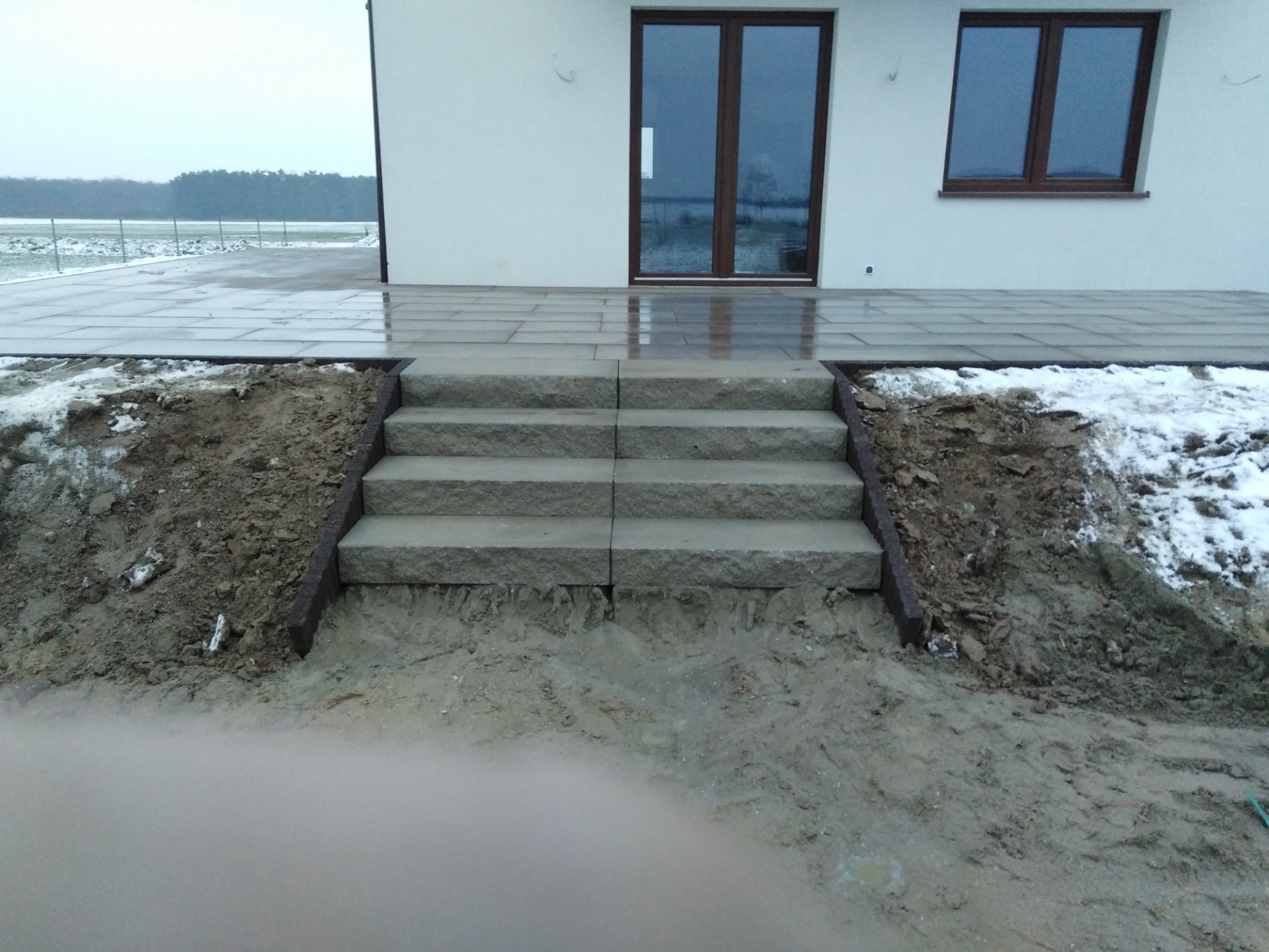 Kamienne schody prowadzące na taras z płytek brukowych przy białym domu w pochmurny dzień.