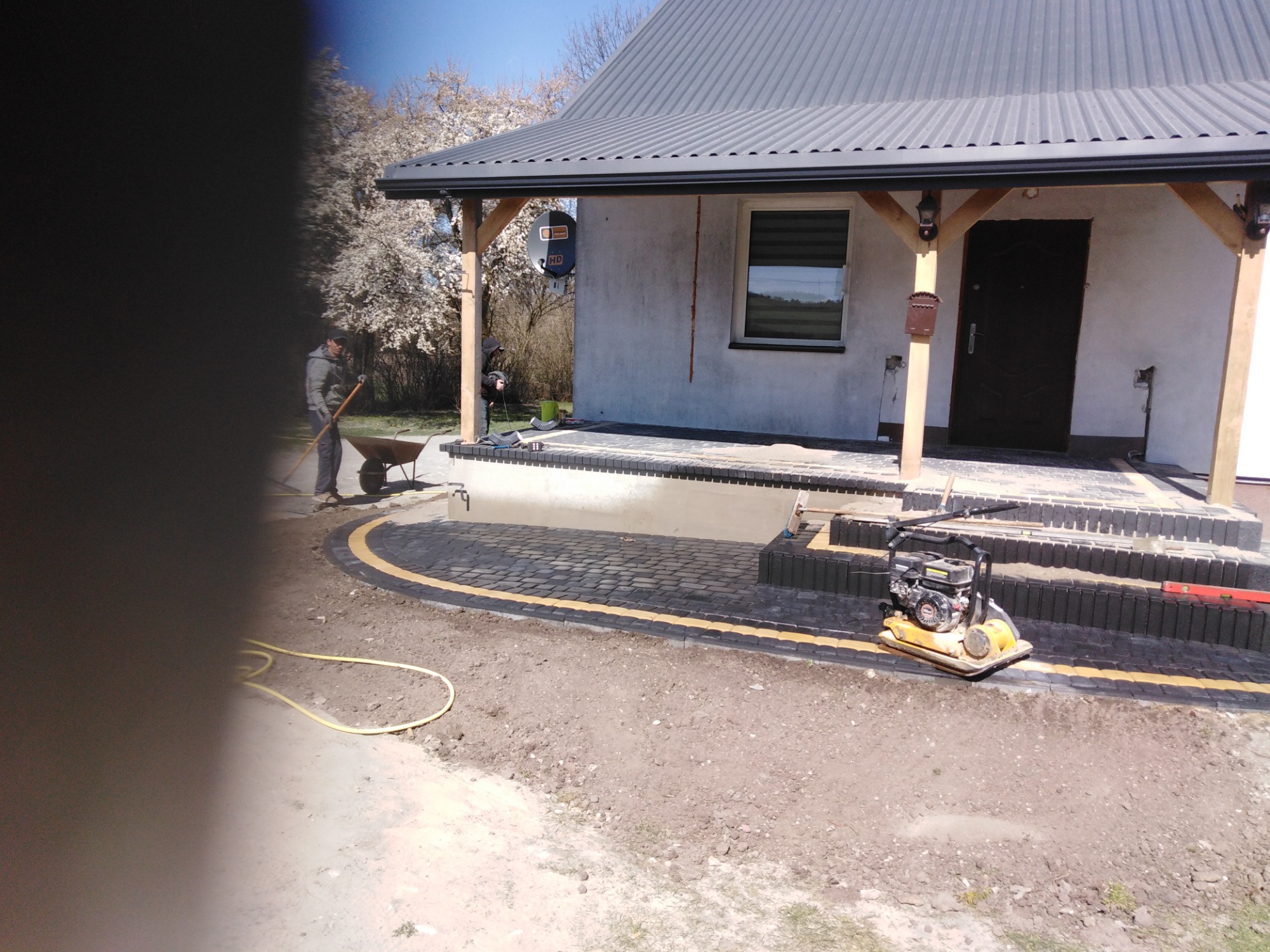 Prace brukarskie przed domem: układanie ciemnej kostki z żółtą obwódką, ubijarka wibracyjna stoi obok, pracownik wyrównuje teren, w tle kwitnące drzewa.