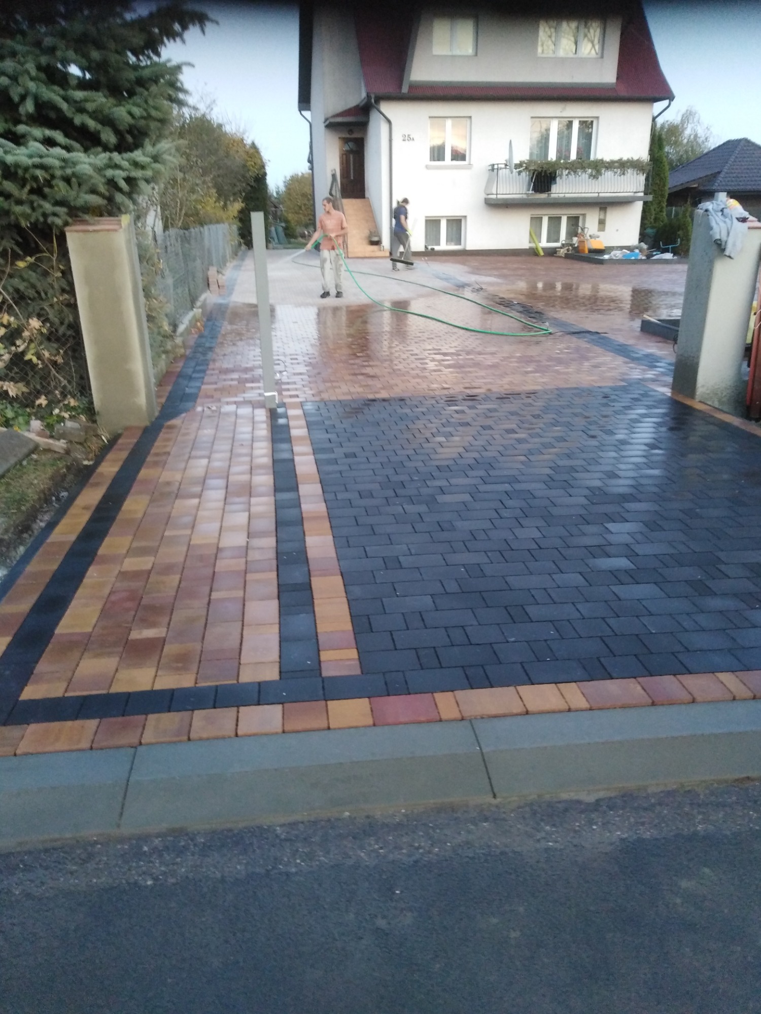 Umyta, dwukolorowa nawierzchnia z kostki brukowej w Kraśniczynie, z widocznym wężem ogrodowym i pracownikami przy domu.