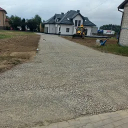 Żwirowa droga do nowego, białego domu z ciemnym dachem, z pracującym traktorem i zagęszczarką w tle, oraz osobą wyrównującą teren grabiami.