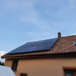 Panele fotowoltaiczne zamontowane na dachu domu z dachówką bitumiczną, widoczne okno dachowe i kominy, błękitne niebo w tle.