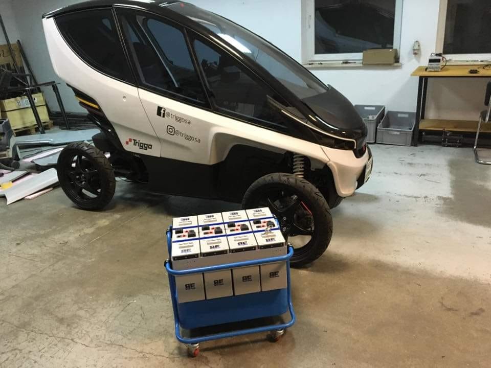 Prototyp trójkołowego pojazdu elektrycznego Triggo z wózkiem akumulatorów, widoczne logo i adresy mediów społecznościowych na karoserii.