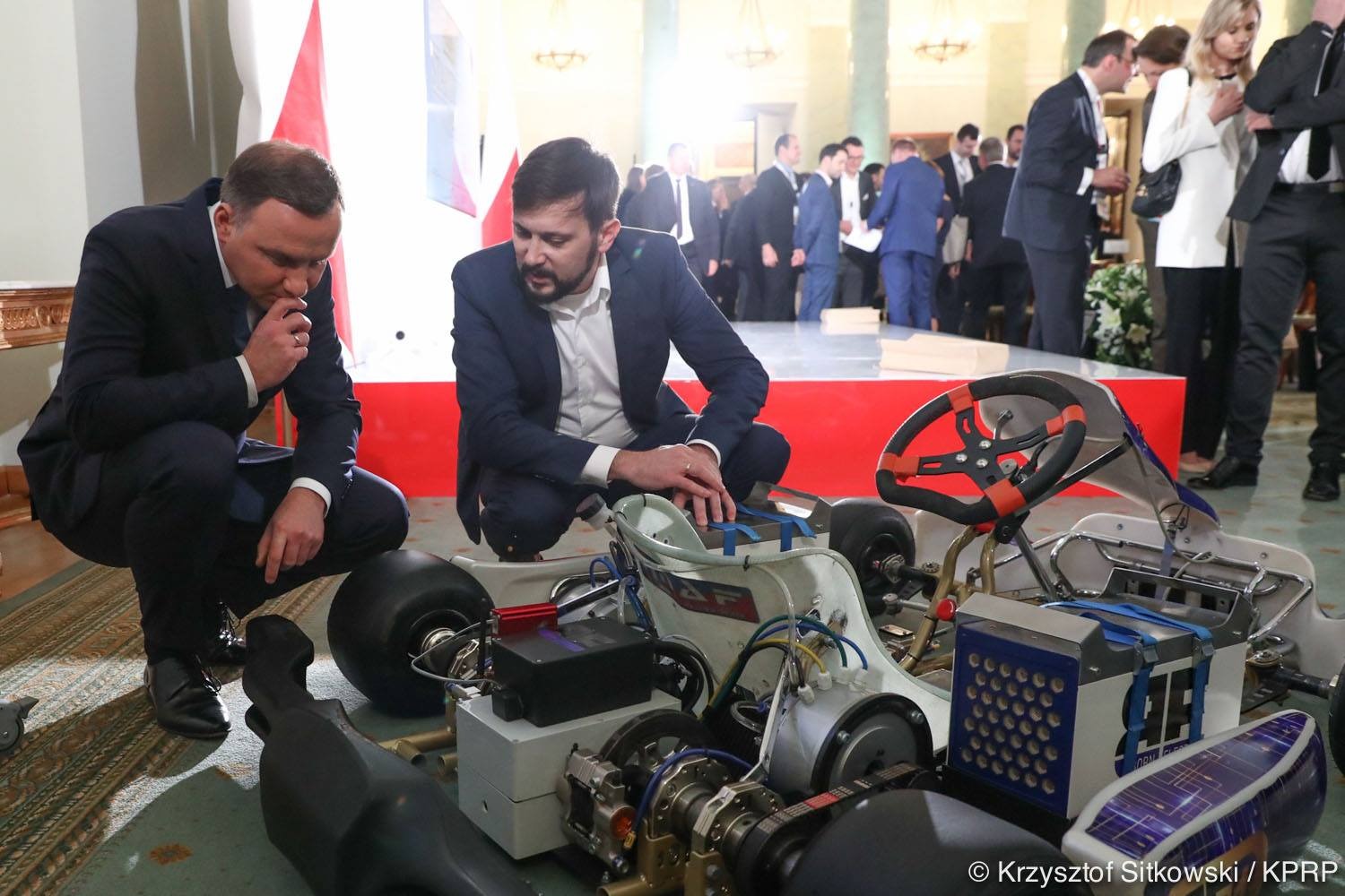 Dwóch mężczyzn w garniturach przykuca nad prototypem gokarta elektrycznego z widocznymi elementami konstrukcyjnymi i elektronicznymi, w tle grupa osób w formalnych strojach.
