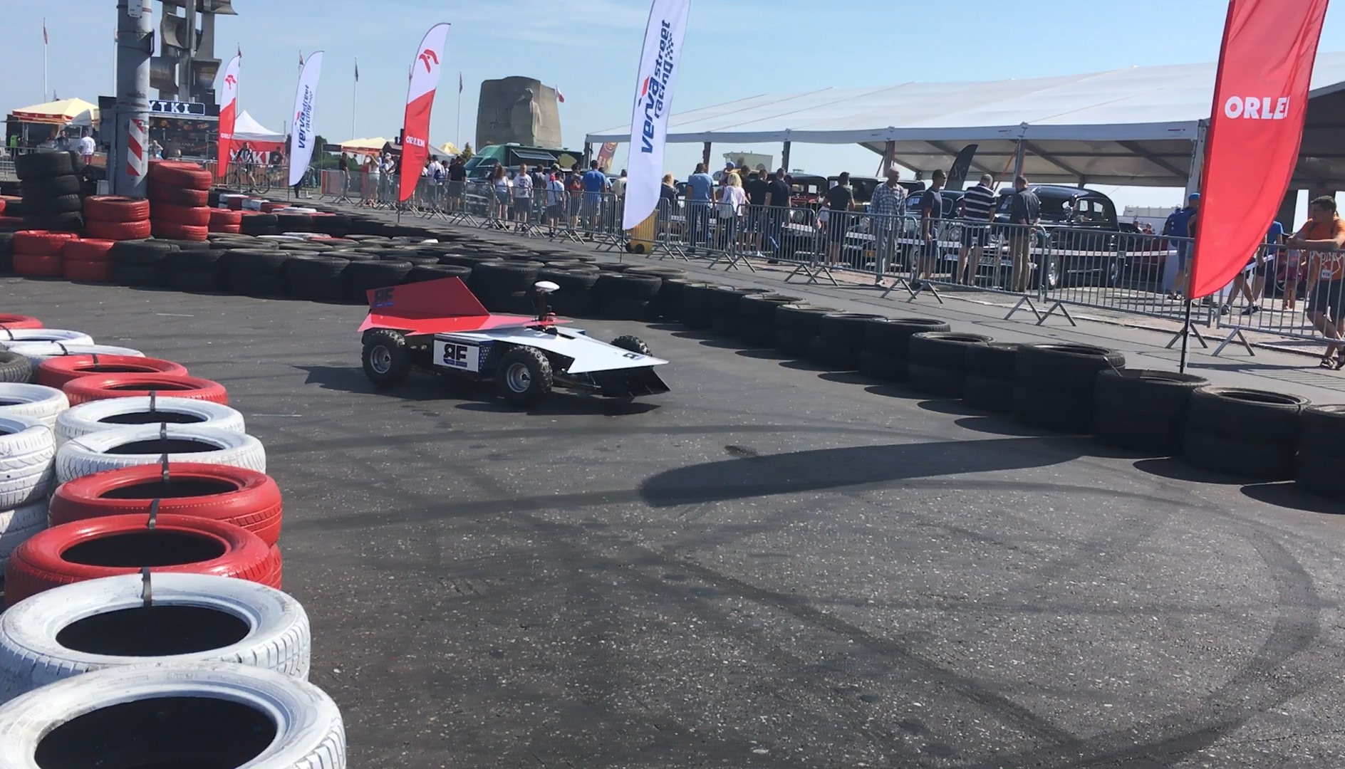 Zdalnie sterowany model bolidu wyścigowego na torze otoczonym oponami, w tle widoczni ludzie i flagi sponsorskie.