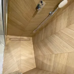 Kabina prysznicowa z wykończeniem drewnopodobnym w jodełkę, nowoczesna armatura, odpływ liniowy w podłodze.