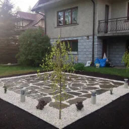 Wykończony fragment ogrodu z kwadratowym patio z nieregularnych kamieni otoczonych białym grysem, z centralnie posadzonym młodym drzewkiem i lampami ogrodowymi.