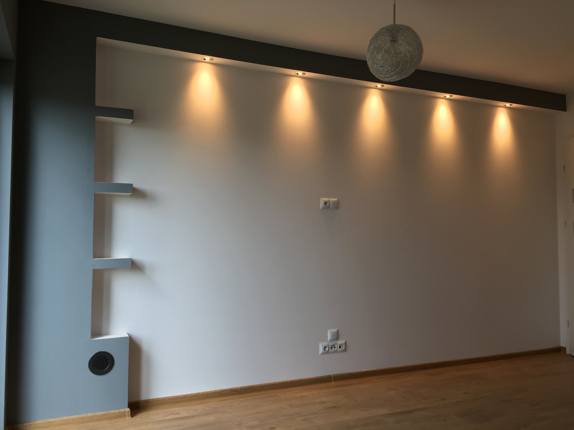 Pusty pokój po remoncie z drewnianą podłogą, białą ścianą z szarymi wnękami i półkami, oświetleniem punktowym w suficie podwieszanym i wiszącą lampą z abażurem z nici.