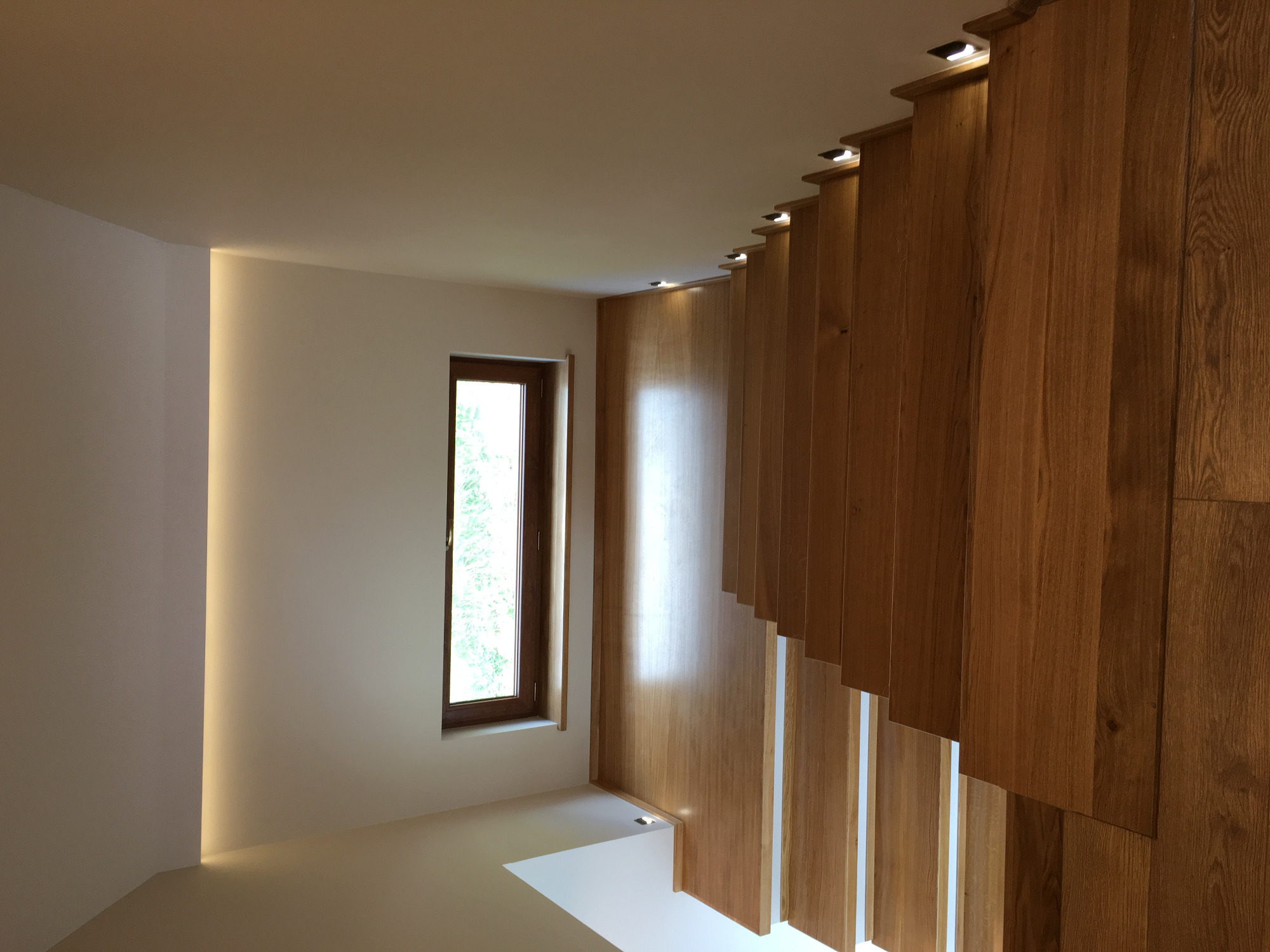 Nowoczesne wnętrze z drewnianą ścianą działową, oświetleniem LED w listwach przypodłogowych i punktowym na górze ściany, oraz oknem z widokiem na zieleń.