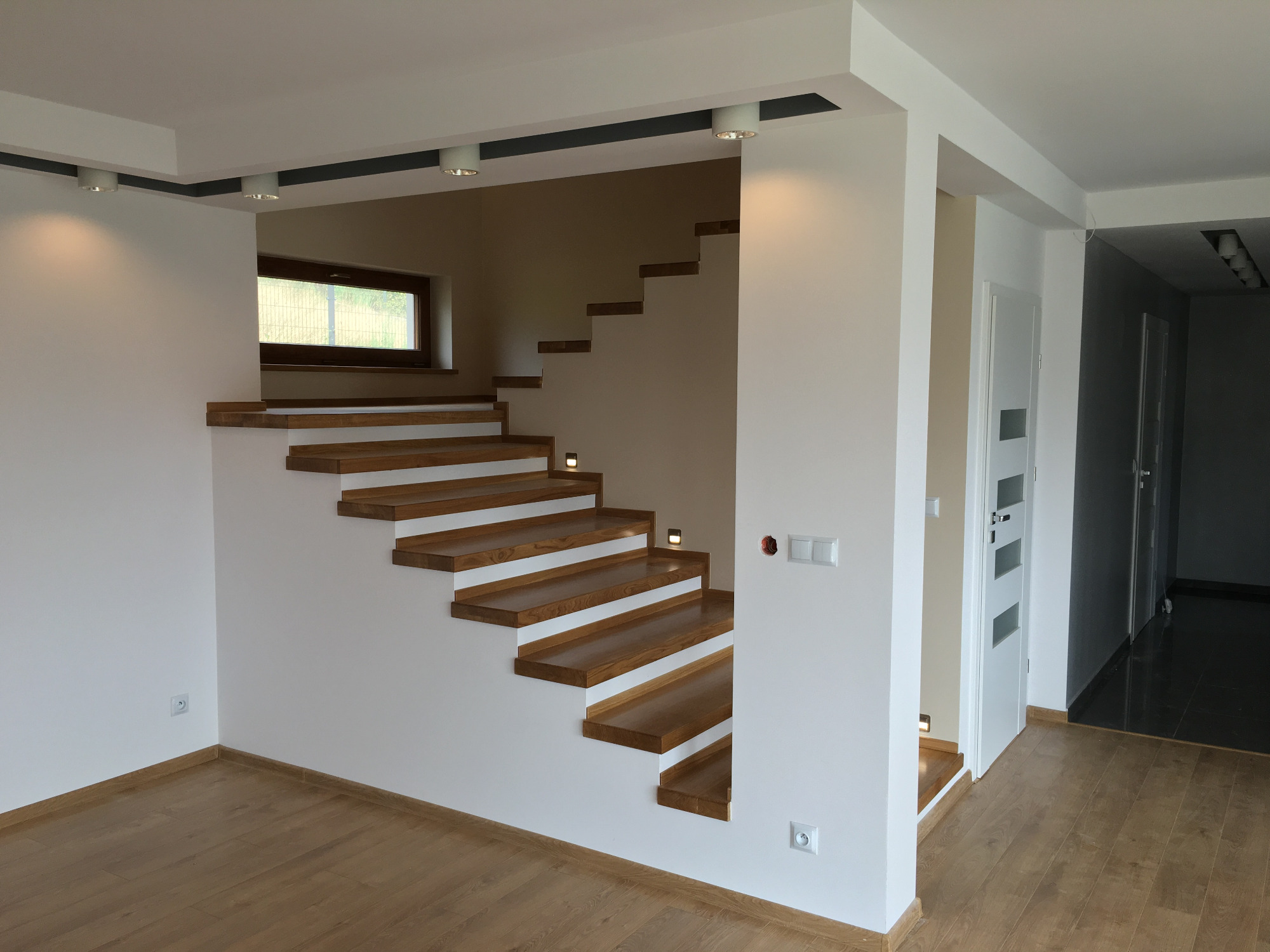 Nowoczesne drewniane schody wspornikowe z oświetleniem punktowym w minimalistycznym wnętrzu, widoczne gładkie, białe ściany i drewniana podłoga.