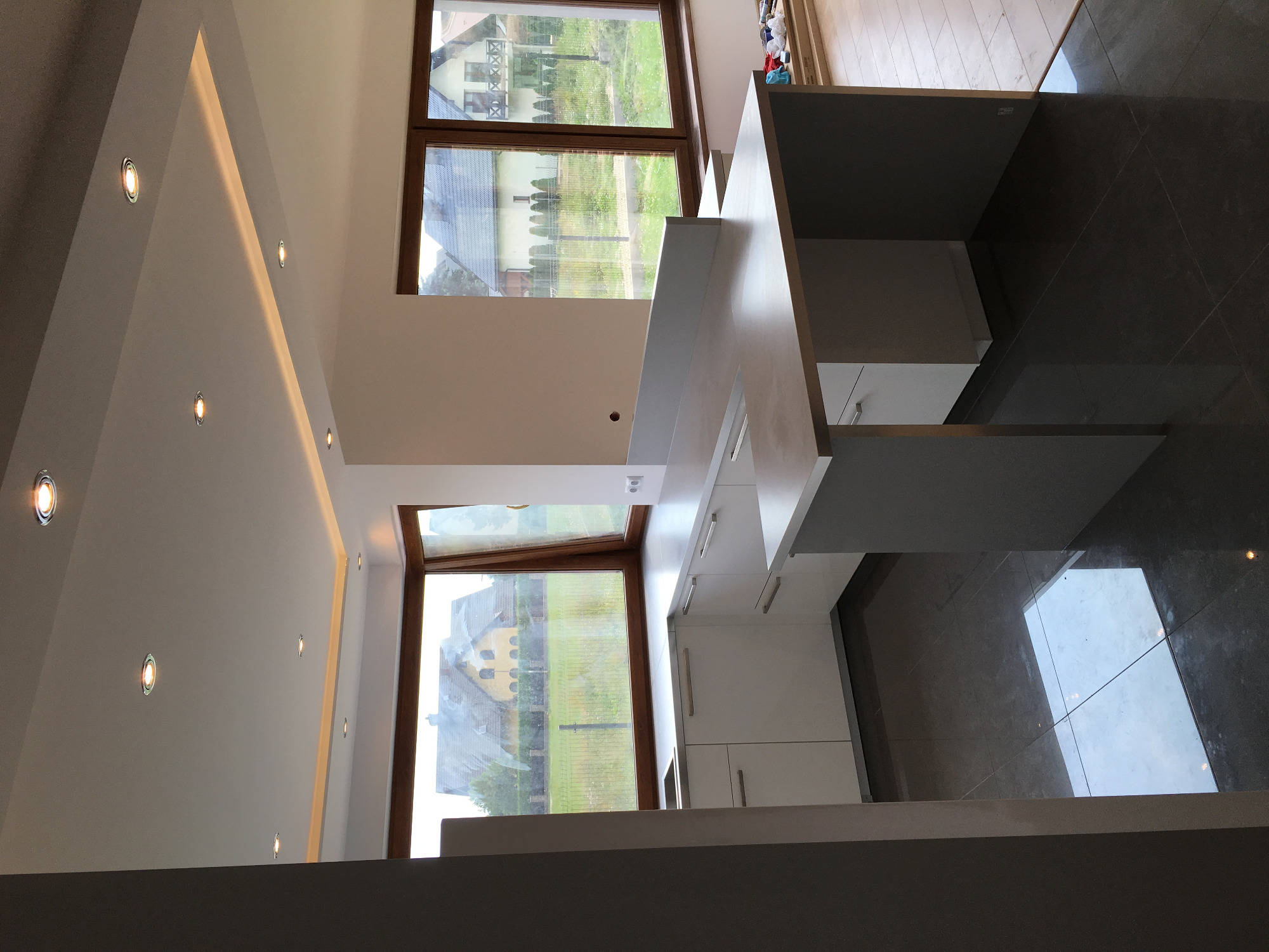 Wnętrze kuchni w trakcie wykańczania, widoczne białe szafki kuchenne, szare płytki podłogowe i duże okna z widokiem na domy i zieleń, oświetlenie punktowe w suficie.