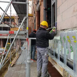 Montaż okna w surowym stanie budynku: pracownik w żółtym kasku uszczelnia ramę okienną silikonem na wysokości rusztowania, widoczne ocieplenie budynku i fragment dźwigu.