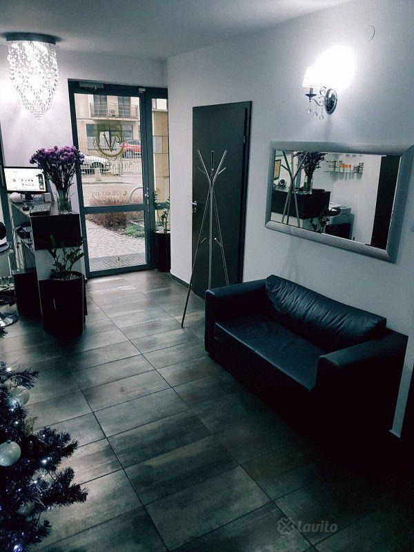 Wnętrze gabinetu z czarną skórzaną sofą, w tle recepcja z fioletowymi kwiatami i widok na ulicę przez szklane drzwi, oświetlone ozdobnym żyrandolem i kinkietem.