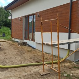 marek1234 - Elewacja domu z drewnopodobną okładziną, fragment rusztowania i wąż do ocieplania. Widoczne okno i fragment dachu. Prace w toku, teren wokół domu przygotowany.