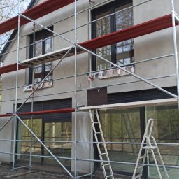 marek1234 - Dom jednorodzinny w trakcie tynkowania elewacji, widoczne rusztowania i drabiny. Świeży tynk w szarym odcieniu. Okna z czarnymi ramami.