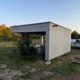 marek1234 - Nowoczesna wiata garażowa z tynkowaną elewacją w trakcie budowy, widoczne materiały budowlane i otoczenie działki. Elementy wykończeniowe w kolorze szarym.