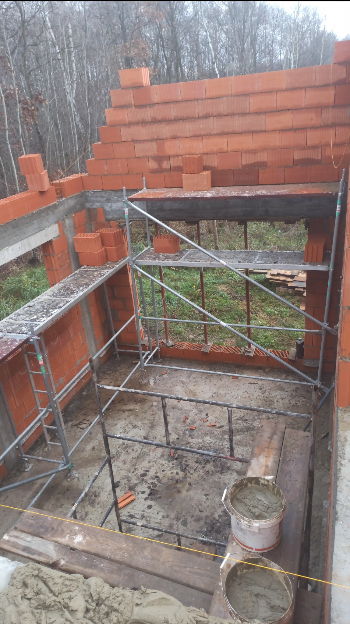 Wznoszenie ścian z czerwonej cegły na budowie domu. Widoczne rusztowania, zaprawa murarska i fragment lasu w tle. Prace murarskie w toku.