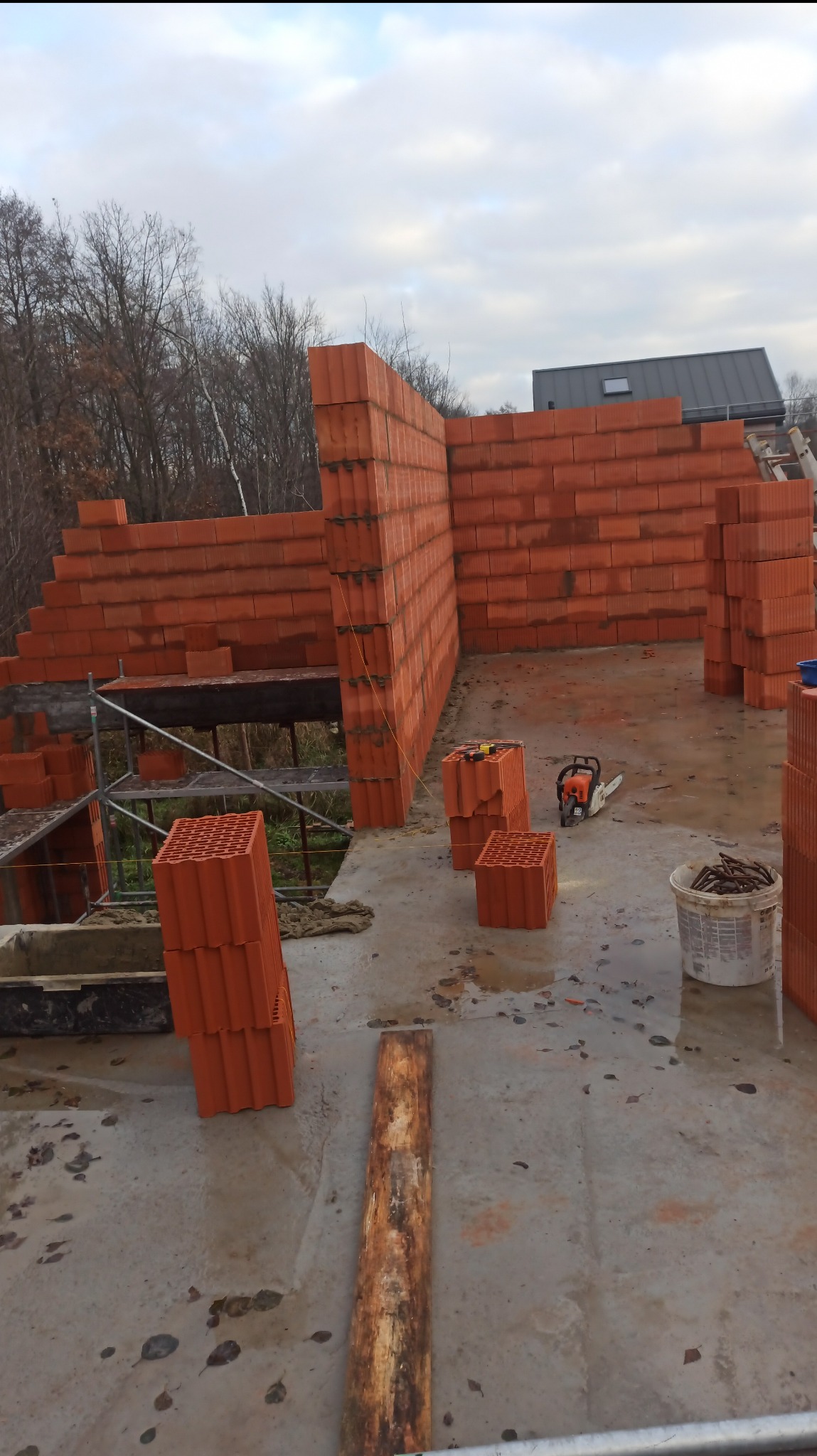 Wznoszenie ścian z czerwonej ceramiki porotherm na budowie domu. Widoczne rusztowania, narzędzia i materiały budowlane. Ujęcie z góry, w tle drzewa i fragment dachu.