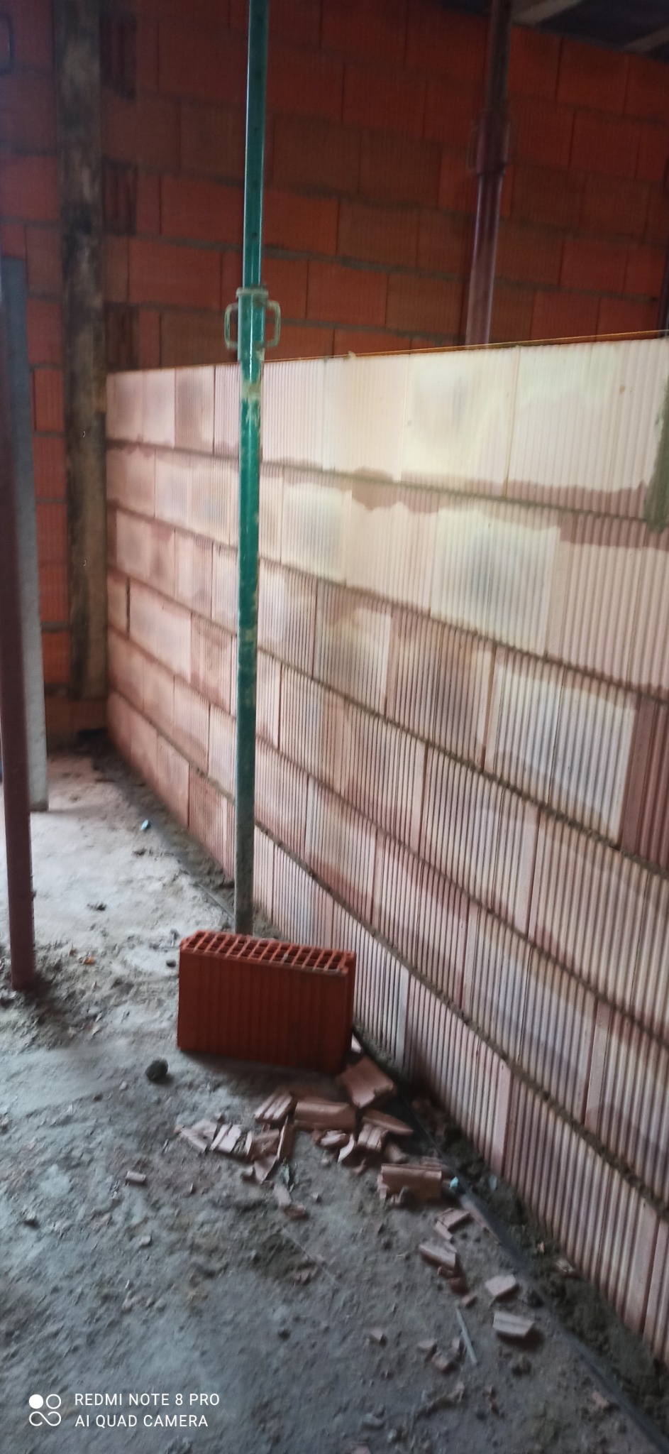 Ściana z pustaków ceramicznych Porotherm w trakcie budowy, podparta zielonym stemplem budowlanym. Na podłodze gruz i fragmenty cegieł. Widok z perspektywy podłogi.