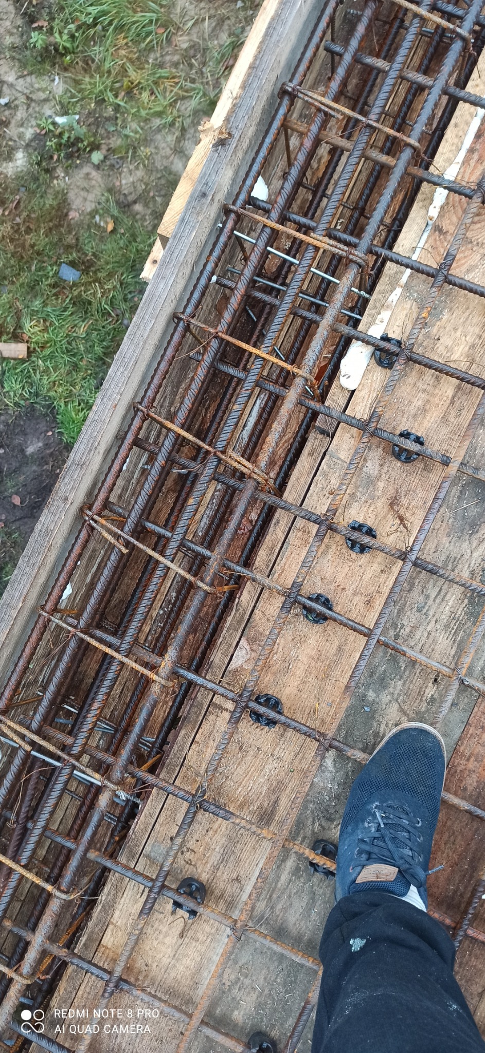 Zbrojenie fundamentów z użyciem dystansów, widoczne drewniane szalunki i fragment buta pracownika. Realizacja z bliska, z perspektywy osoby stojącej na konstrukcji.