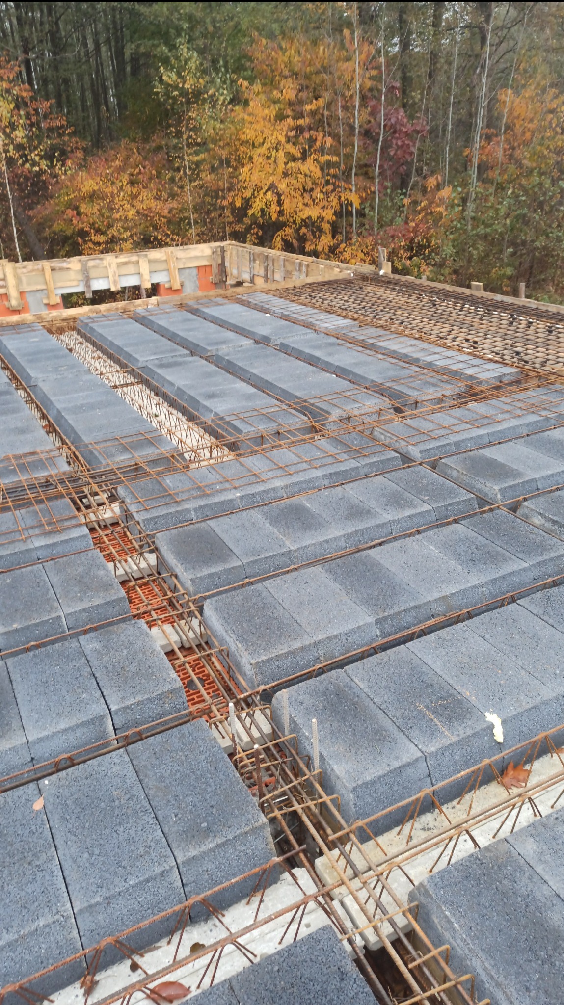 Szare bloczki stropowe ułożone na konstrukcji zbrojeniowej, widoczne elementy szalunku i jesienne drzewa w tle. Etap budowy domu.