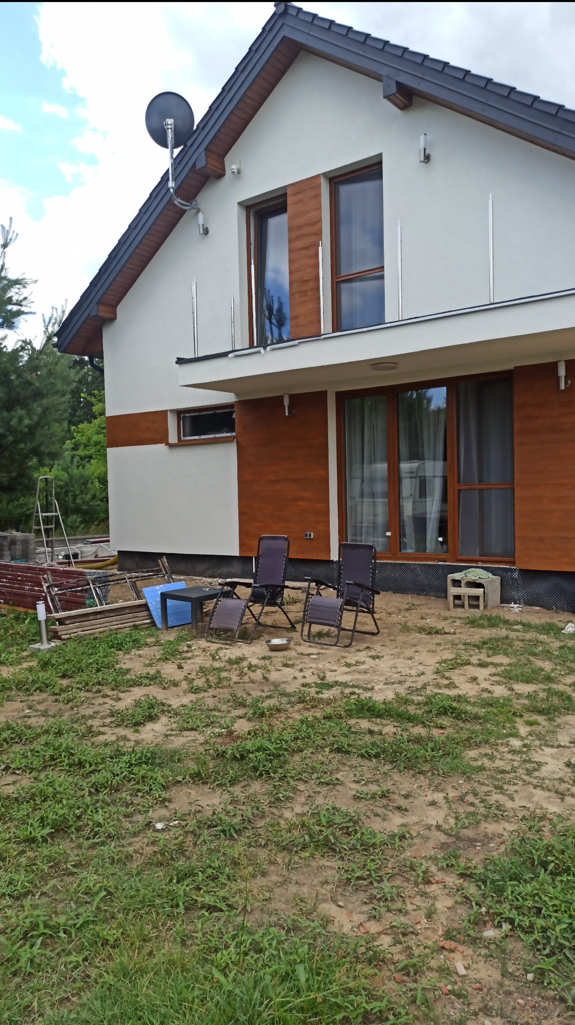 Dwukondygnacyjny dom z jasną elewacją i drewnianą okładziną. Widoczne okna z brązowymi ramami, balkon z metalową balustradą i antena satelitarna na dachu. Na trawniku krzesła.