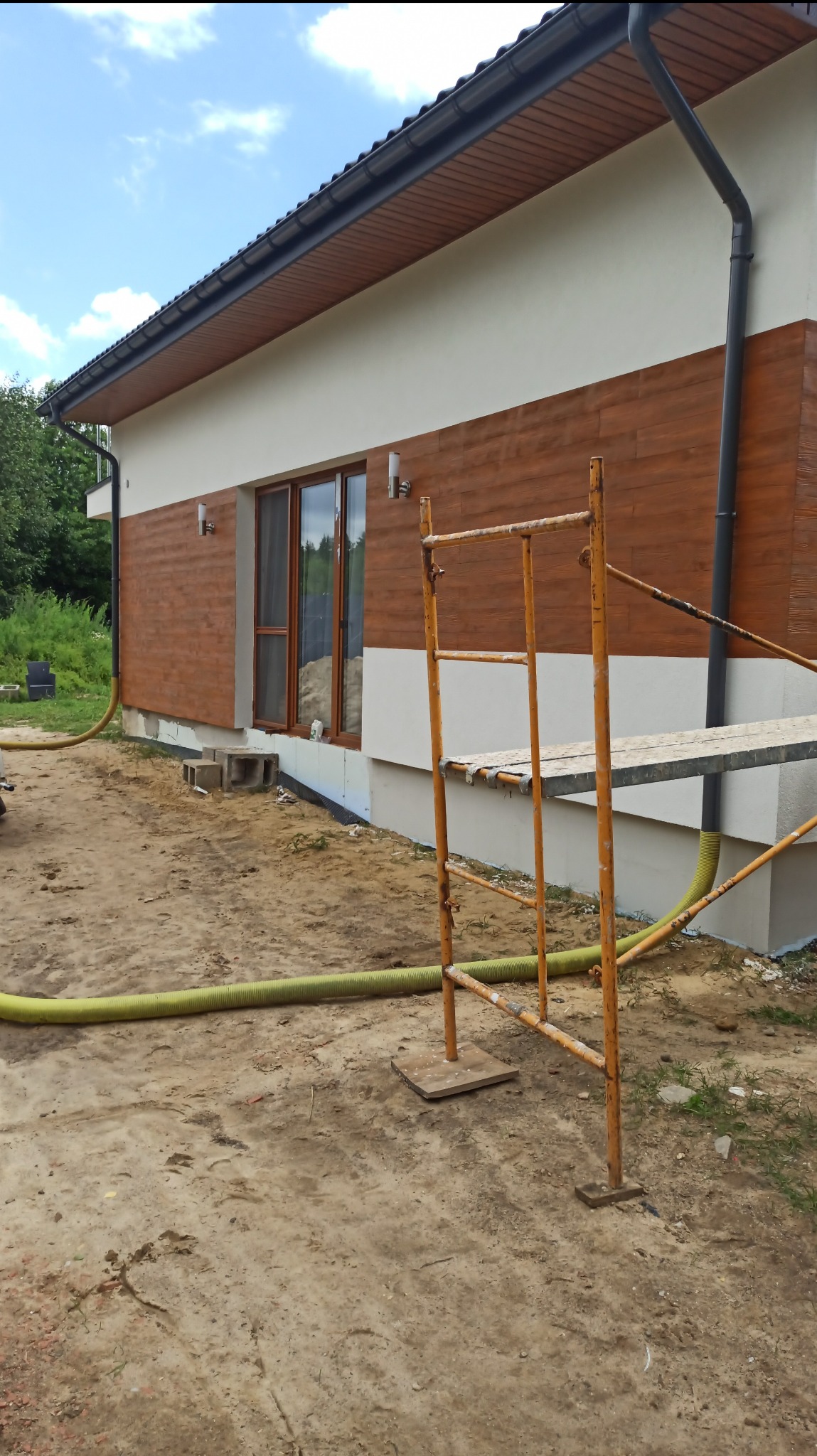 Elewacja domu z drewnopodobną okładziną, fragment rusztowania i wąż do ocieplania. Widoczne okno i fragment dachu. Prace w toku, teren wokół domu przygotowany.