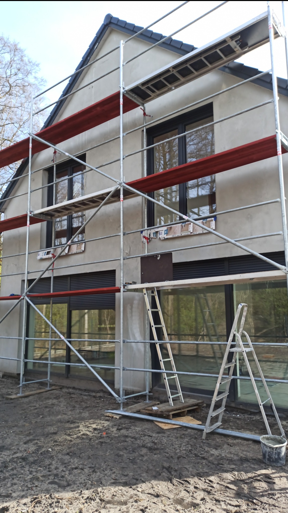 Dom jednorodzinny w trakcie tynkowania elewacji, widoczne rusztowania i drabiny. Świeży tynk w szarym odcieniu. Okna z czarnymi ramami.