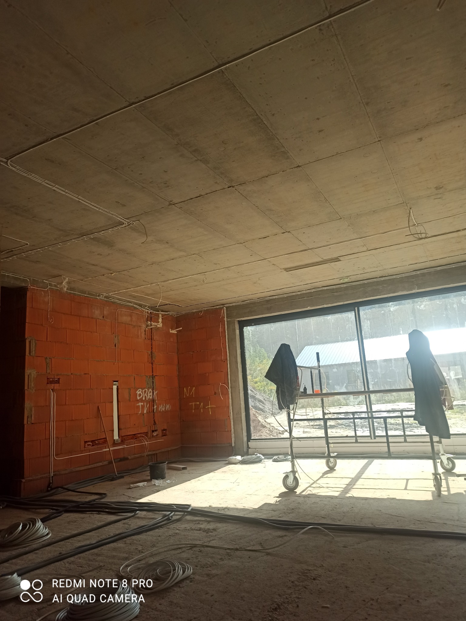 Surowy stan wnętrza domu w trakcie budowy: widoczne instalacje elektryczne na ceglanej ścianie, betonowy sufit i duże okno z widokiem na zewnątrz.