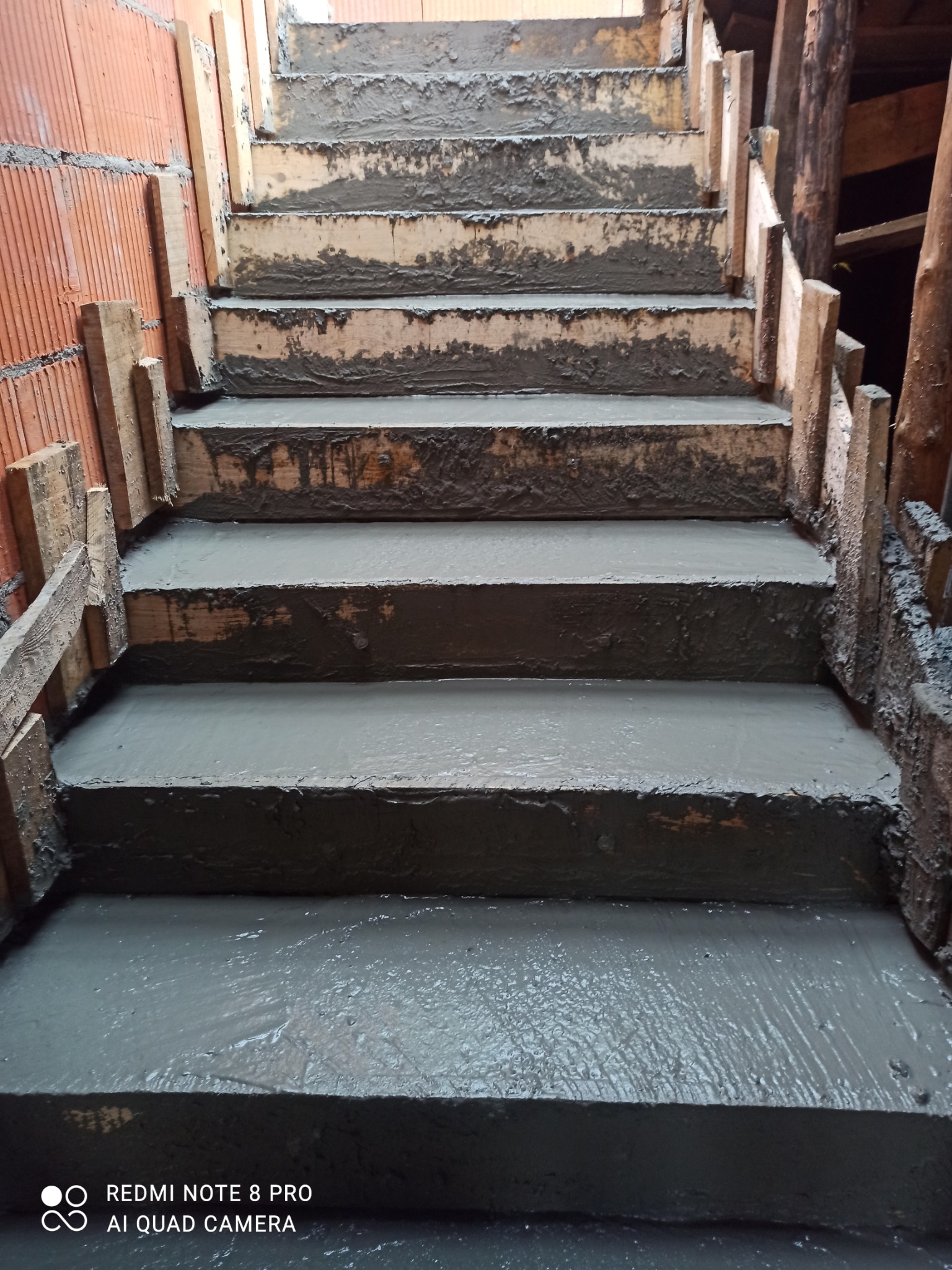 Wylewanie betonowych schodów w drewnianym szalunku, widok z dołu. Świeży beton na stopniach, ceglana ściana z boku. Etap budowy schodów.