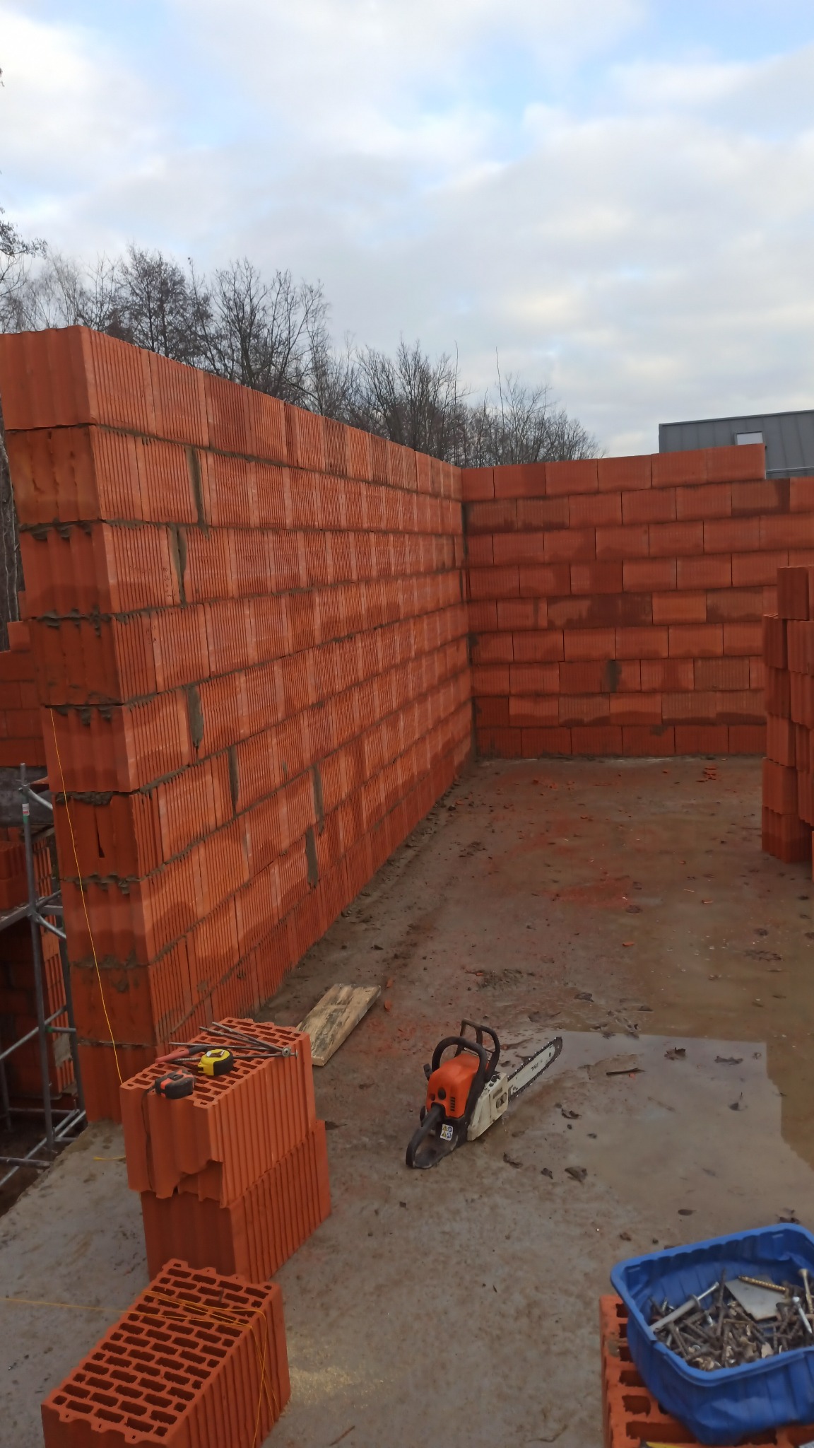 Wzniesiona ściana z czerwonych bloczków ceramicznych na mokrej posadzce. Obok leżą narzędzia: piła spalinowa, miara i poziomica. Widoczne rusztowanie i niebo.