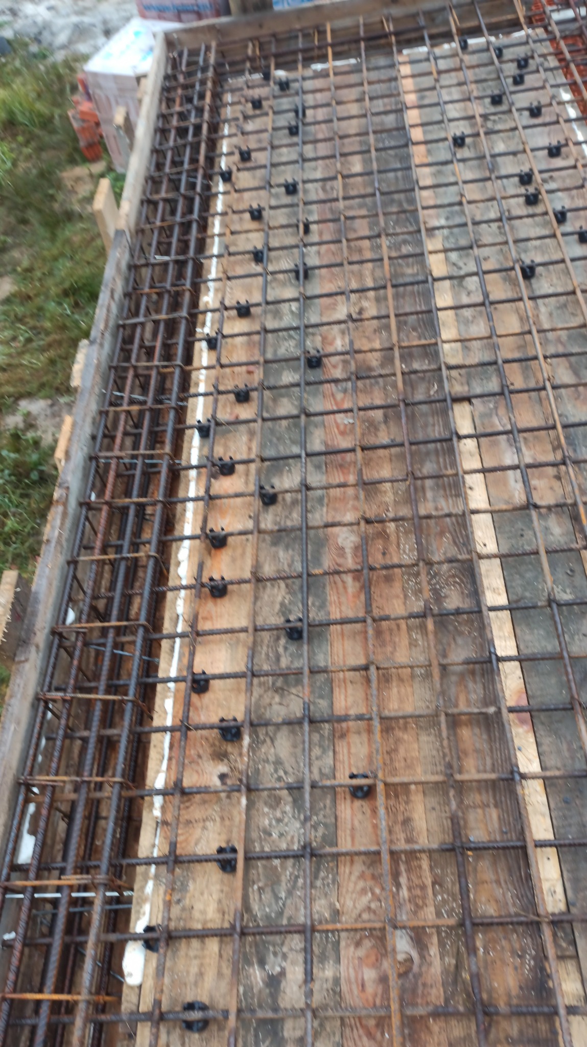 Zbrojenie fundamentu domu jednorodzinnego, widok z góry na siatkę z prętów stalowych, drewniane szalunki i elementy dystansowe. Etap budowy.