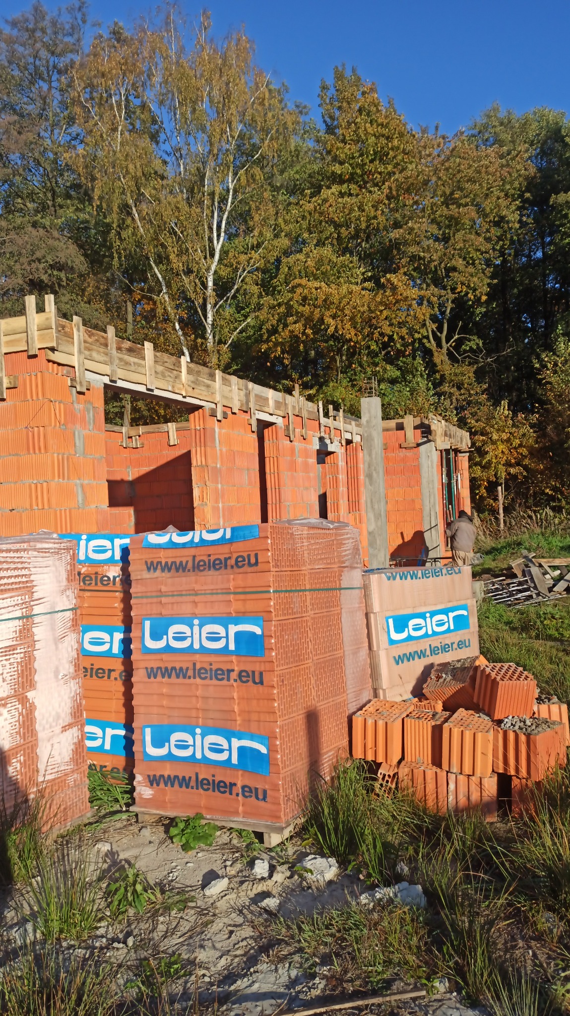 Wznoszenie ścian domu z czerwonej cegły Leier, widoczne palety z cegłami, drewniane szalunki i jesienne drzewa w tle pod błękitnym niebem. Etap budowy domu.