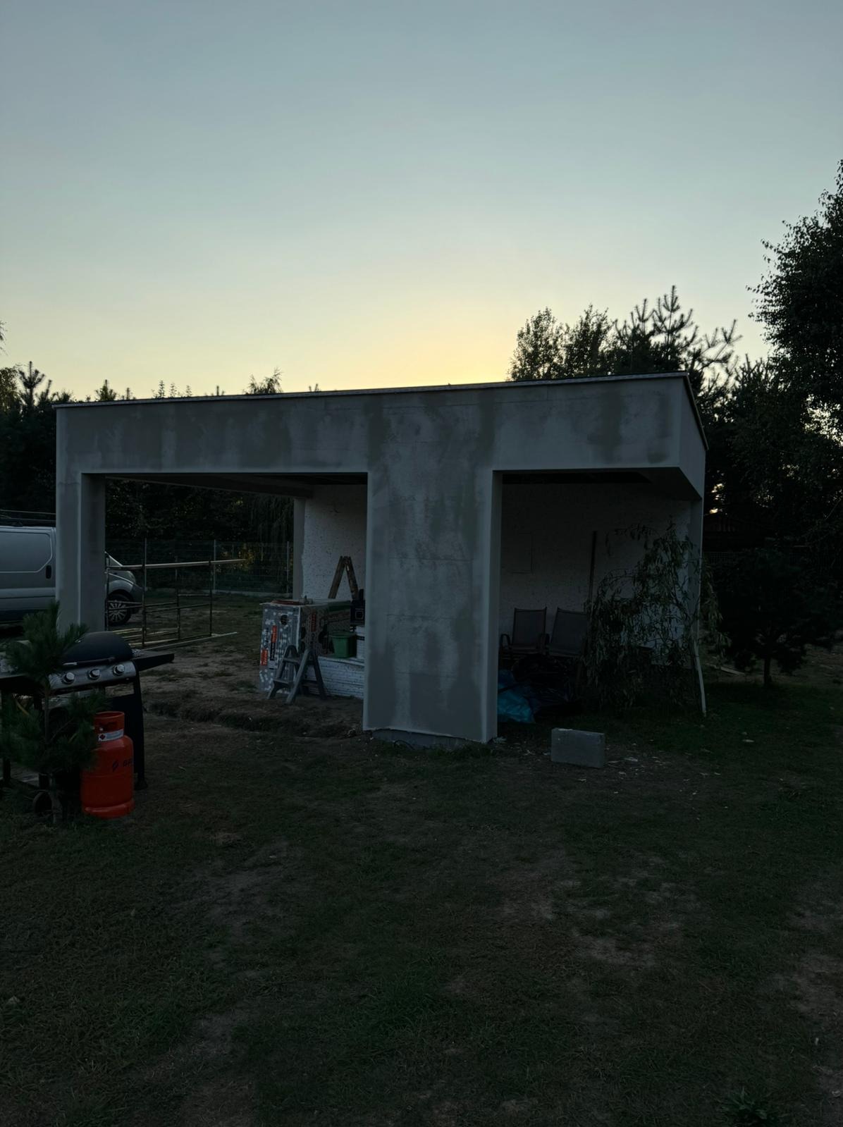 Surowy, otynkowany garaż/wiata z dwoma wjazdami, w tle zieleń i zachodzące słońce. Na pierwszym planie grill i butla gazowa. Widoczne narzędzia i drabina.