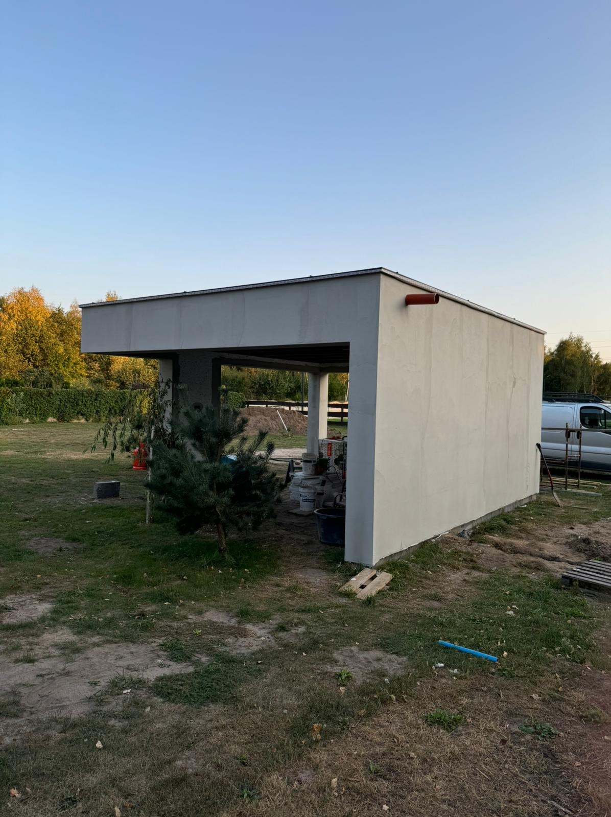 Nowoczesna wiata garażowa z tynkowaną elewacją w trakcie budowy, widoczne materiały budowlane i otoczenie działki. Elementy wykończeniowe w kolorze szarym.