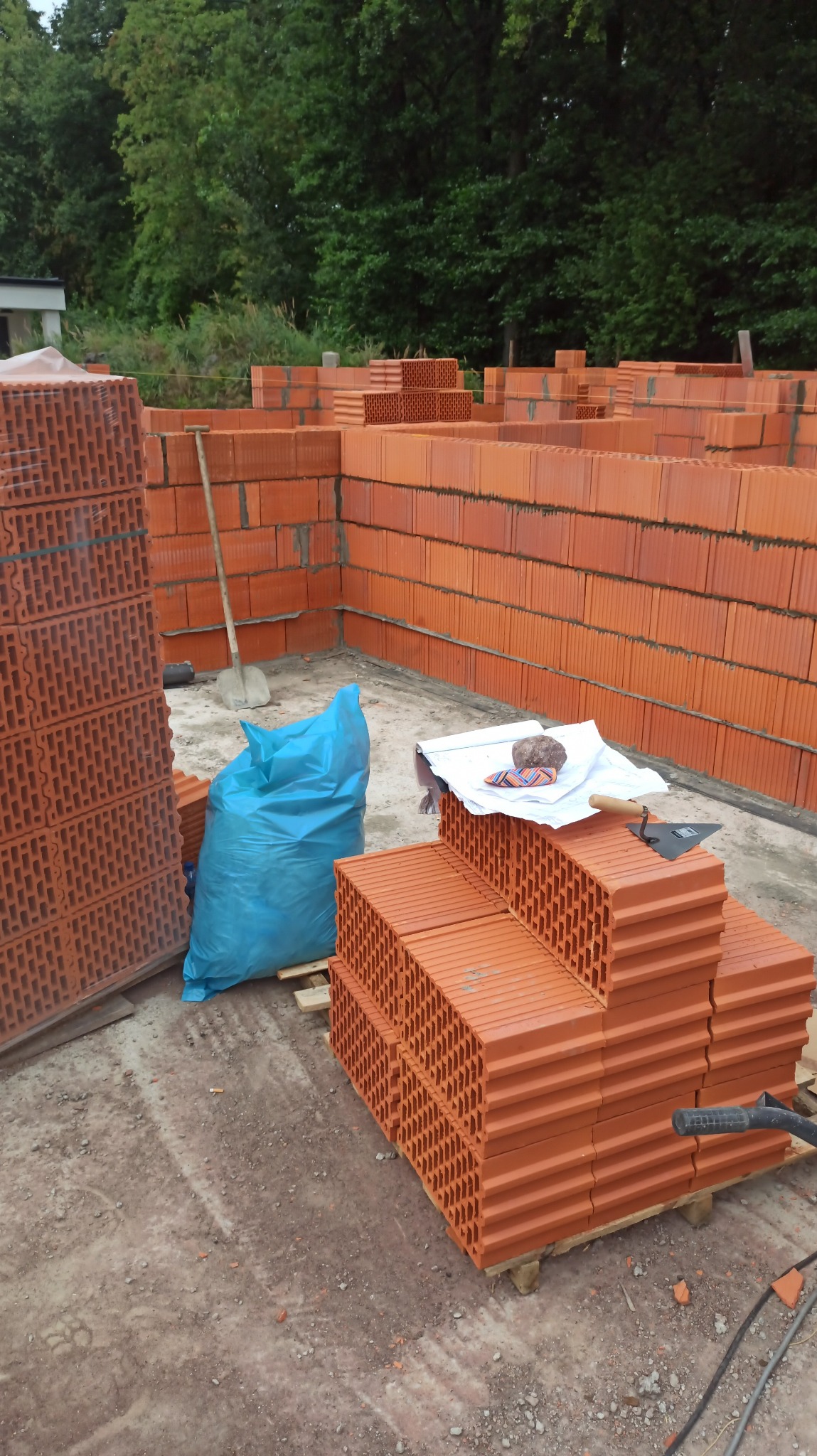 Wznoszenie ścian z czerwonej cegły ceramicznej. Częściowo wymurowane ściany, palety z cegłami, narzędzia murarskie i niebieski worek na placu budowy.