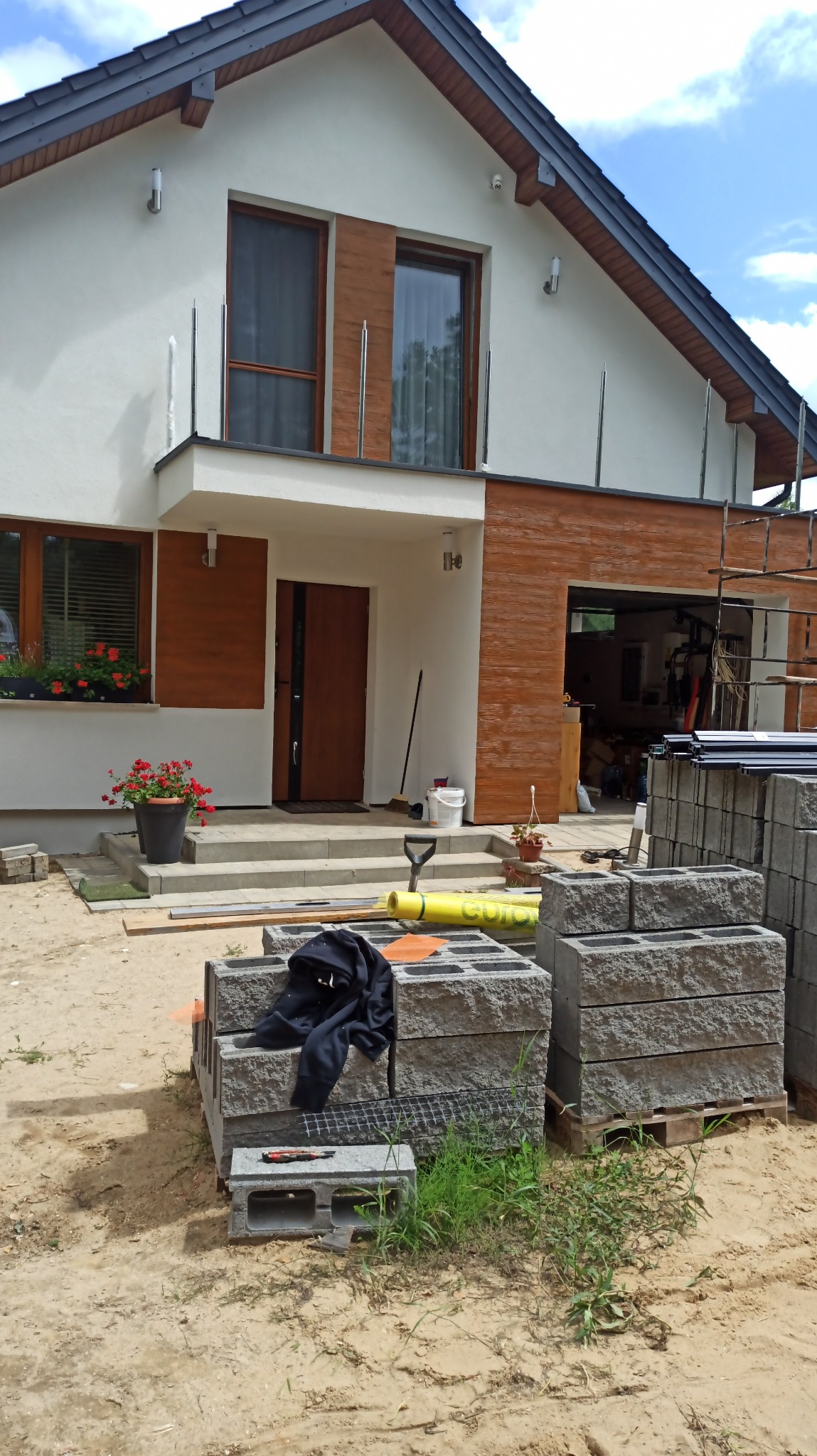 Nowoczesny dom jednorodzinny w trakcie budowy. Widoczne bloczki betonowe, narzędzia i fragment elewacji z drewnianym akcentem. Ujęcie z poziomu gruntu.