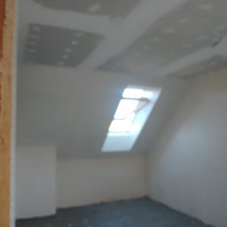 SlaBud - Poddasze w trakcie prac wykończeniowych: widoczne płyty kartonowo-gipsowe na skosach sufitu, okno dachowe i przygotowana podłoga. Ściany w trakcie szpachlowania, widoczne niedoskonałości. Zdjęcie...