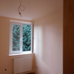 SlaBud - Pokój po malowaniu na jasny odcień, widoczne świeżo pomalowane ściany, białe okno z widokiem na zieleń, grzejnik i drewniana podłoga; brak wykończenia wokół grzejnika.