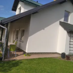 Boczny widok dwukondygnacyjnego domu z białą elewacją i zielonym dachem, z widocznym fragmentem tarasu z meblami ogrodowymi i zadbanym trawnikiem.