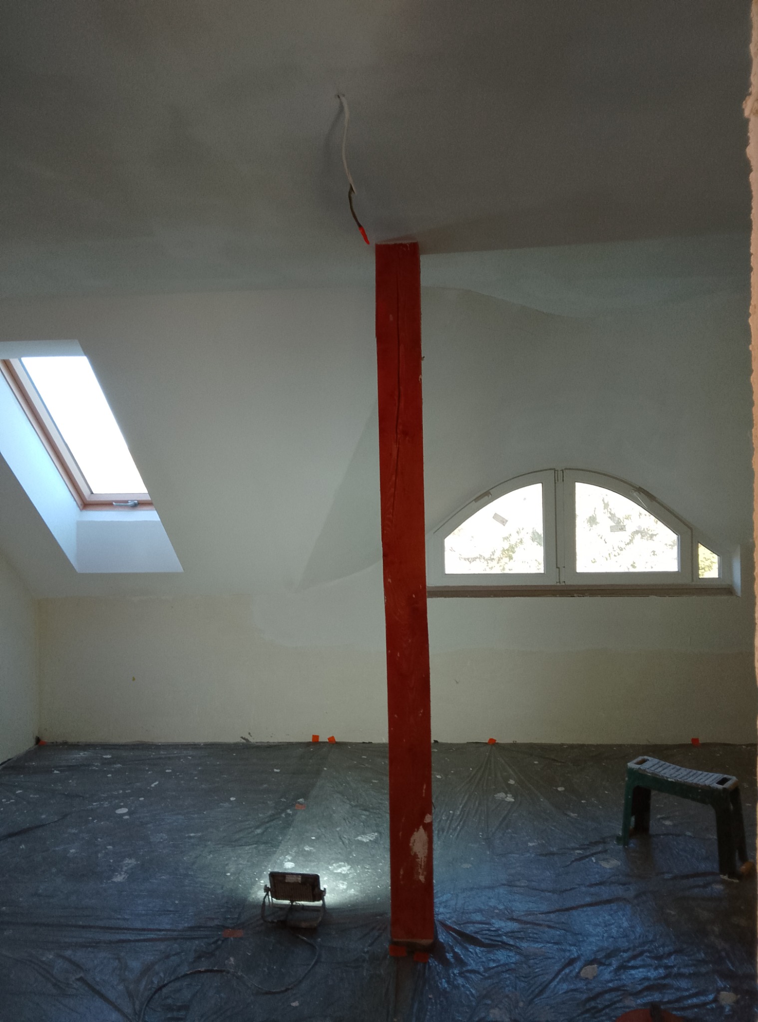 Poddasze w trakcie prac wykończeniowych: zabezpieczona folią podłoga, oświetlenie robocze, drewniany słup pomalowany na czerwono, okno dachowe i okno w szczycie. Widoczne ślady po szpachlowaniu...