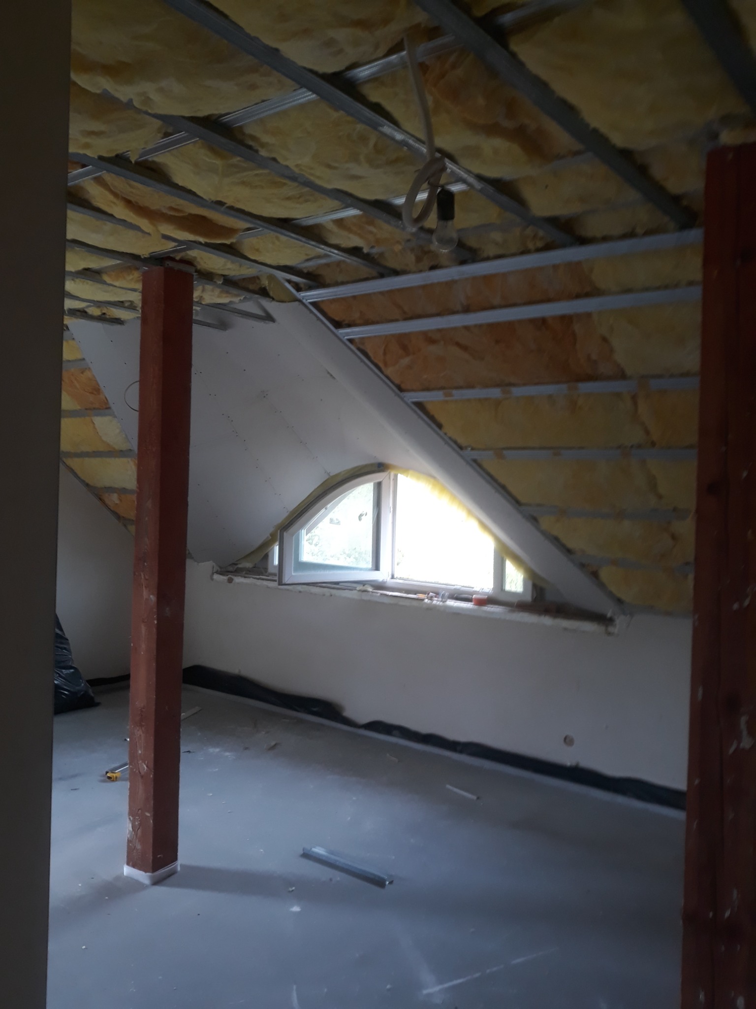 Poddasze w trakcie adaptacji: widoczna izolacja z wełny mineralnej na skośnym suficie, metalowy stelaż, częściowo zamontowana płyta karton-gips przy oknie dachowym o łukowym kształcie, surowa...