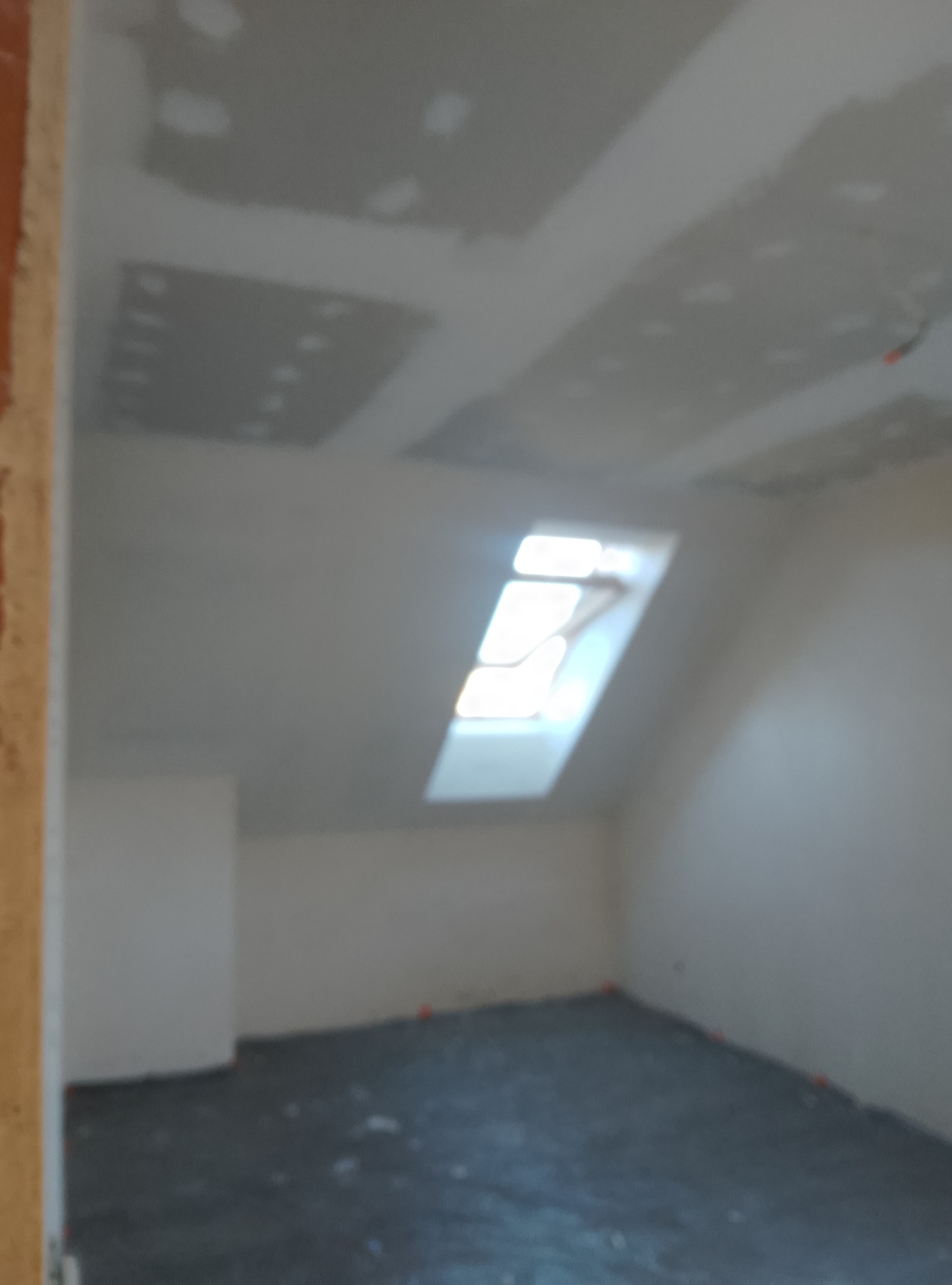 Poddasze w trakcie prac wykończeniowych: widoczne płyty kartonowo-gipsowe na skosach sufitu, okno dachowe i przygotowana podłoga. Ściany w trakcie szpachlowania, widoczne niedoskonałości. Zdjęcie...