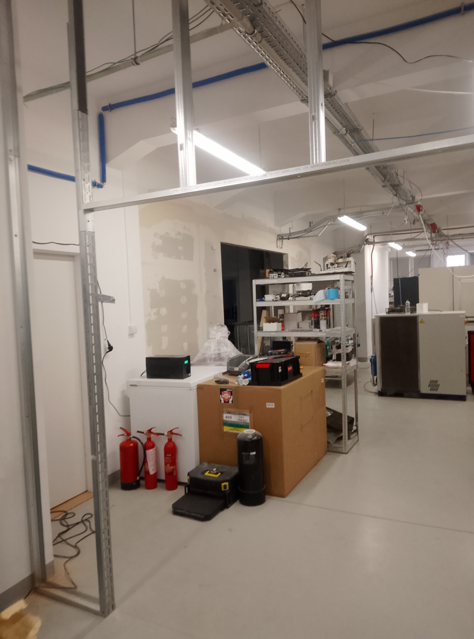 Wnętrze w trakcie prac remontowych, widoczne stalowe profile konstrukcyjne ścian działowych, częściowo zagruntowana ściana, instalacje podwieszane pod sufitem oraz sprzęt i materiały budowlane w tle.