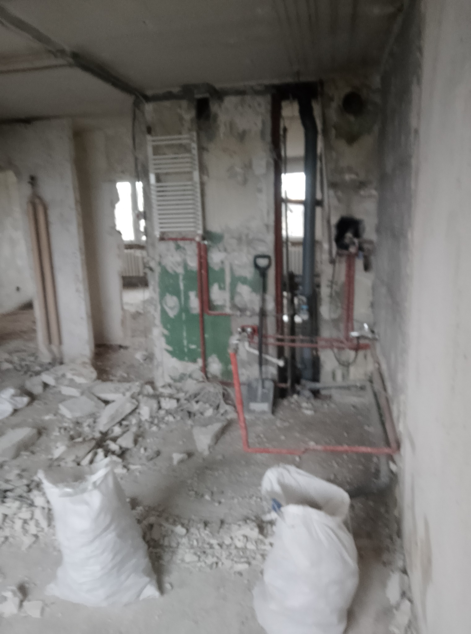 Wnętrze w trakcie remontu: pobojowisko z gruzem, worki z zaprawą, odsłonięte instalacje grzewcze i stara drabinka grzejnikowa na ścianie, widok na sąsiednie pomieszczenie.