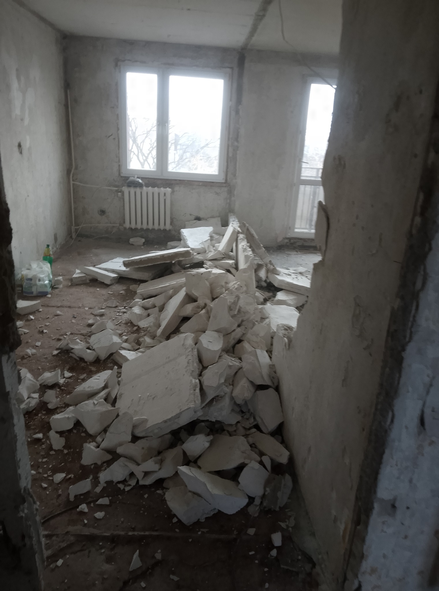 Wnętrze pokoju w trakcie demontażu, widoczne gruzowisko z białych płyt, odsłonięte ściany i okna z widokiem na drzewa.