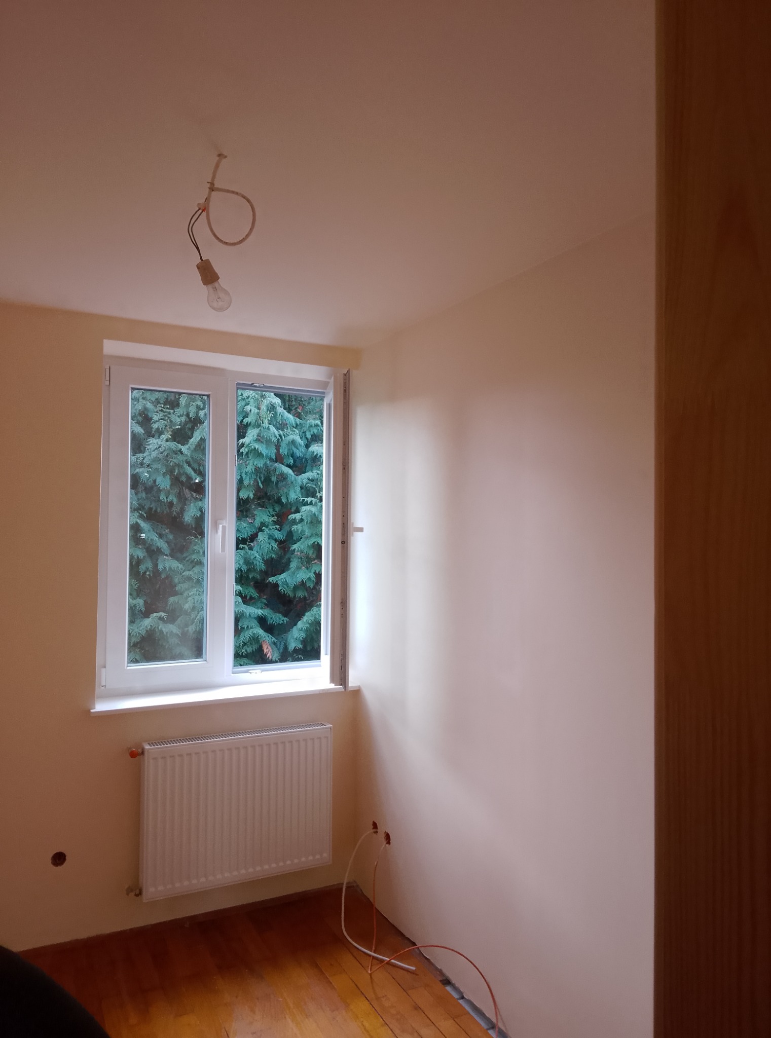 Pokój po malowaniu na jasny odcień, widoczne świeżo pomalowane ściany, białe okno z widokiem na zieleń, grzejnik i drewniana podłoga; brak wykończenia wokół grzejnika.