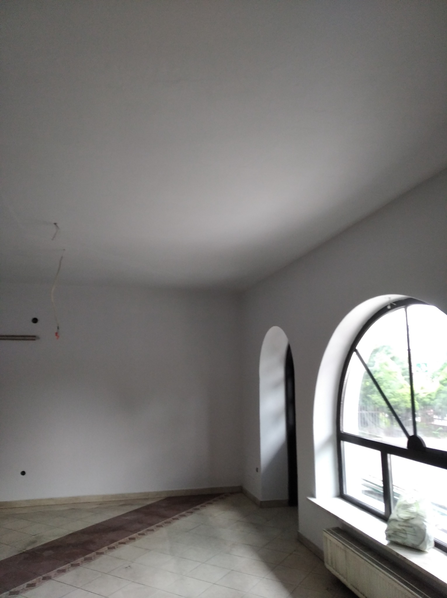 Pusty, jasny pokój po remoncie z dwoma łukowymi oknami, widoczne ślady po instalacjach elektrycznych na ścianie i worki z zaprawą pod oknem.