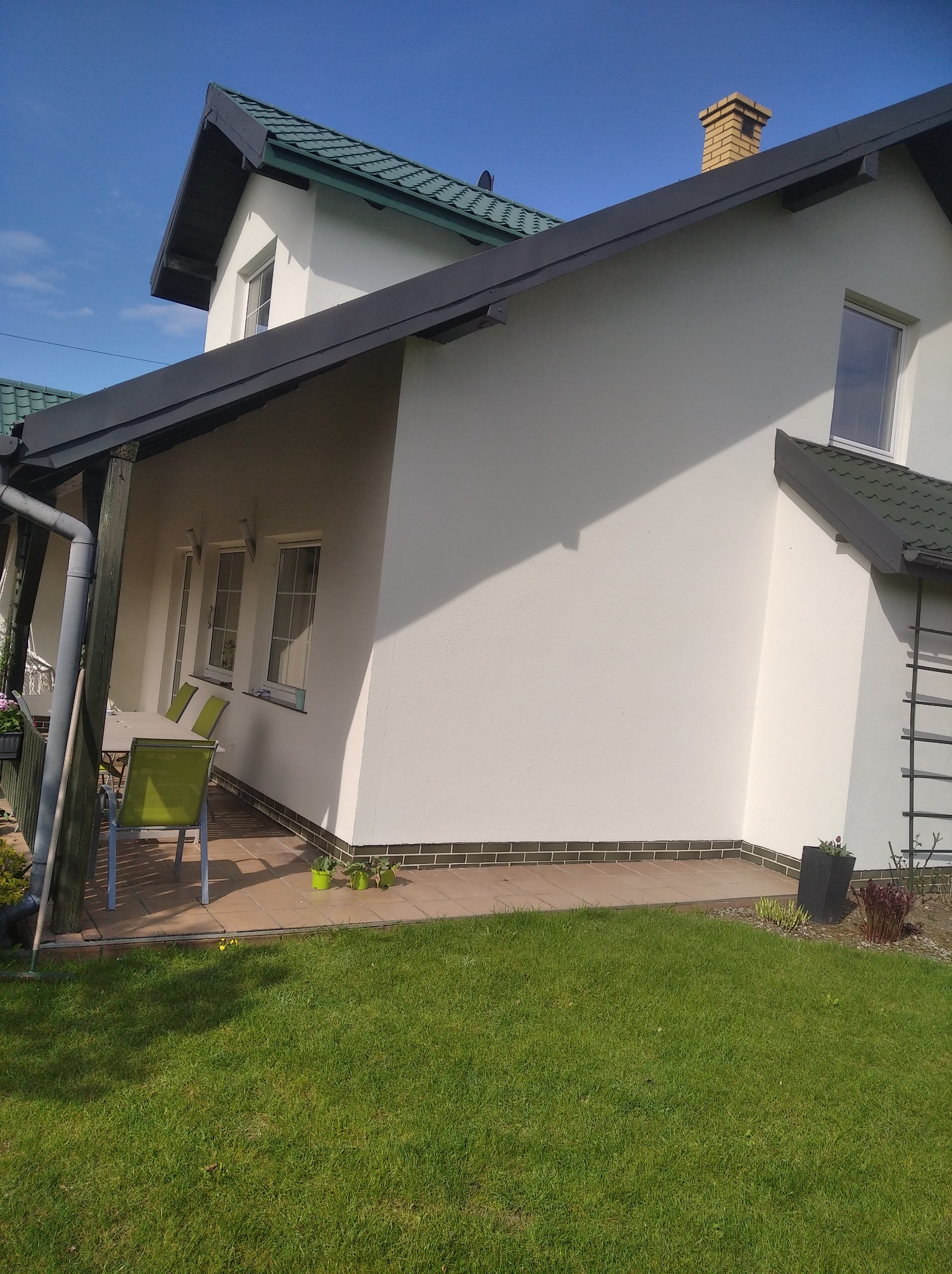 Boczny widok dwukondygnacyjnego domu z białą elewacją i zielonym dachem, z widocznym fragmentem tarasu z meblami ogrodowymi i zadbanym trawnikiem.